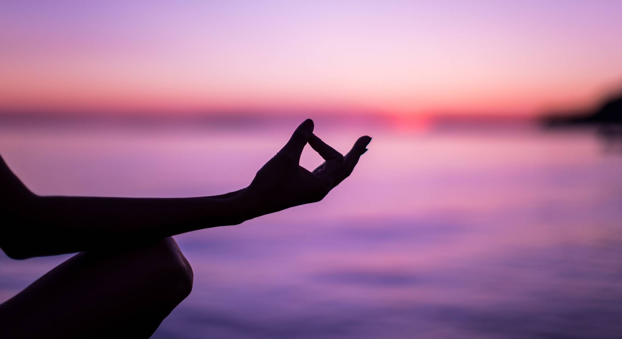 person meditating while a setting sun turns the sky and water a beautiful tint of purple