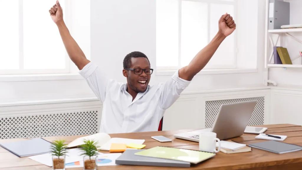 A manager is smiling with their hands up while sitting in front a laptop placed on a desk.